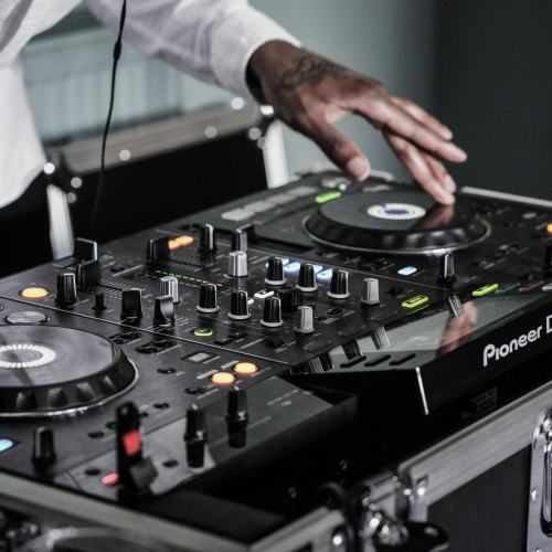 A DJ is using a Pioneer mixer and turntables, adjusting knobs and discs with a hand, wearing a white shirt.