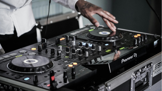 A DJ is using a Pioneer mixer and turntables, adjusting knobs and discs with a hand, wearing a white shirt.