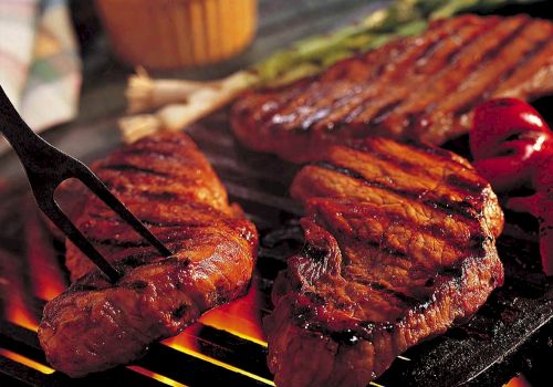 The image shows grilled steaks sizzling over an open flame, with a fork lifting one piece. Roasted red peppers and a yellow sauce are in the background.