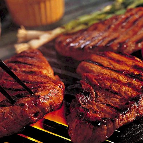 The image shows grilled steaks sizzling over an open flame, with a fork lifting one piece. Roasted red peppers and a yellow sauce are in the background.