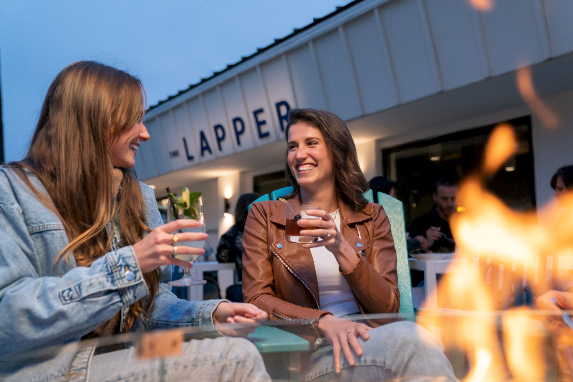 Two women are enjoying drinks and laughing by a fire pit at an outdoor venue with a sign that reads 