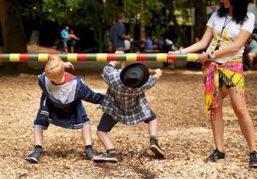 Two children are playing limbo while an adult holds the pole. The scene is set outdoors in a park with a wood-chip ground covering.