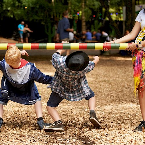 Two children are playing limbo while an adult holds the pole. The scene is set outdoors in a park with a wood-chip ground covering.