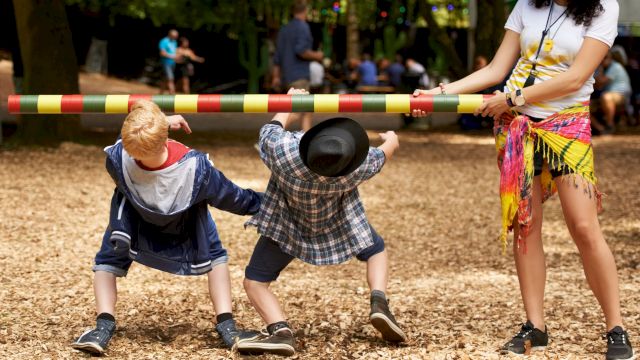 Two children are playing limbo while an adult holds the pole. The scene is set outdoors in a park with a wood-chip ground covering.