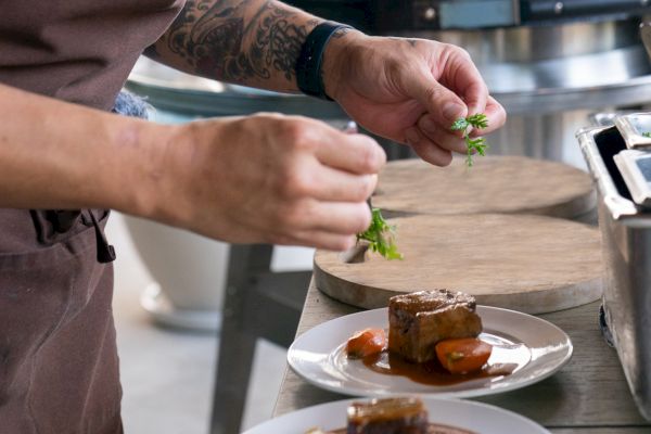 A chef is plating a dish with meat and vegetables, adding garnish to two plates in a kitchen setting.