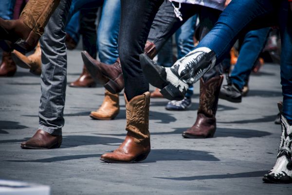 People wearing cowboy boots participating in a group dance, lifting their legs in unison on a concrete surface.