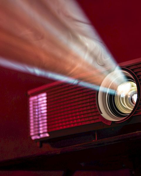 A projector emits a bright beam of light in a dark room, highlighting its lens and creating a vibrant display on a red background.