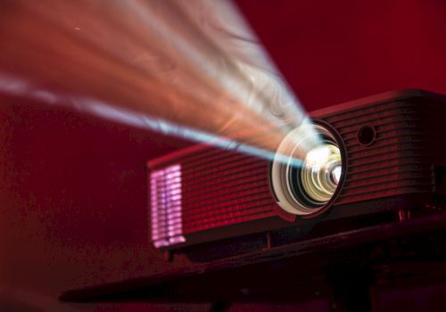 A projector emits a bright beam of light in a dark room, highlighting its lens and creating a vibrant display on a red background.