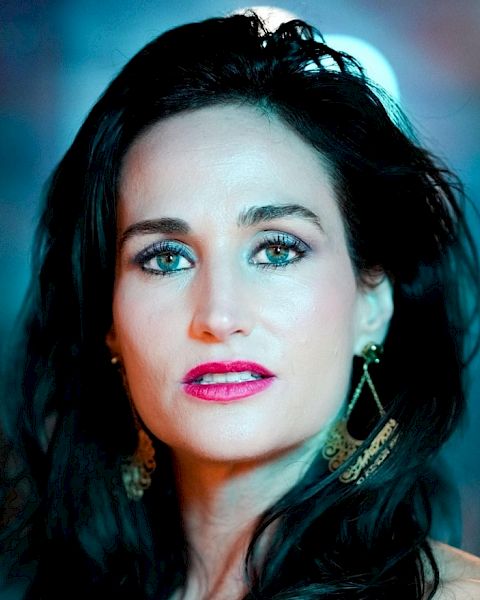 The image shows a woman with long dark hair, wearing gold earrings and makeup, with a blurred background that has blue and orange hues.