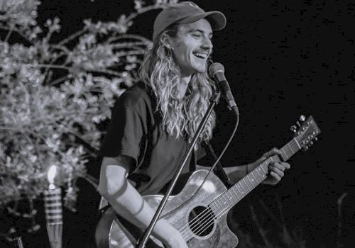 A person with long hair, wearing a cap, is smiling and playing an acoustic guitar while performing on stage, with a microphone in front of them.