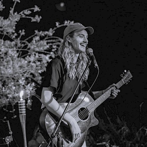 A person with long hair, wearing a cap, is smiling and playing an acoustic guitar while performing on stage, with a microphone in front of them.