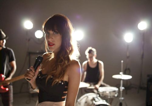 A woman holds a microphone, standing in front of musicians playing drums and guitar, with stage lights shining brightly in the background.