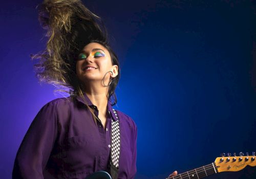 A person with colorful makeup and a purple shirt is playing an electric guitar against a blue and purple background, their hair flying dramatically.