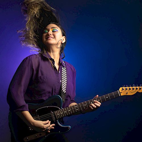 A person with colorful makeup and a purple shirt is playing an electric guitar against a blue and purple background, their hair flying dramatically.