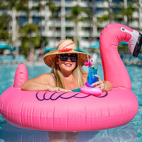 A woman in a sunhat and sunglasses relaxes in a flamingo float in a pool, holding a cocktail with a small flamingo decoration, with buildings behind.