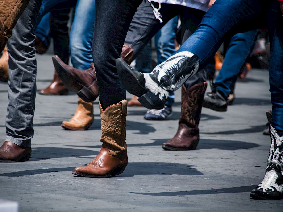 A group of people are wearing cowboy boots and participating in a dance, all in various stages of lifting their legs.