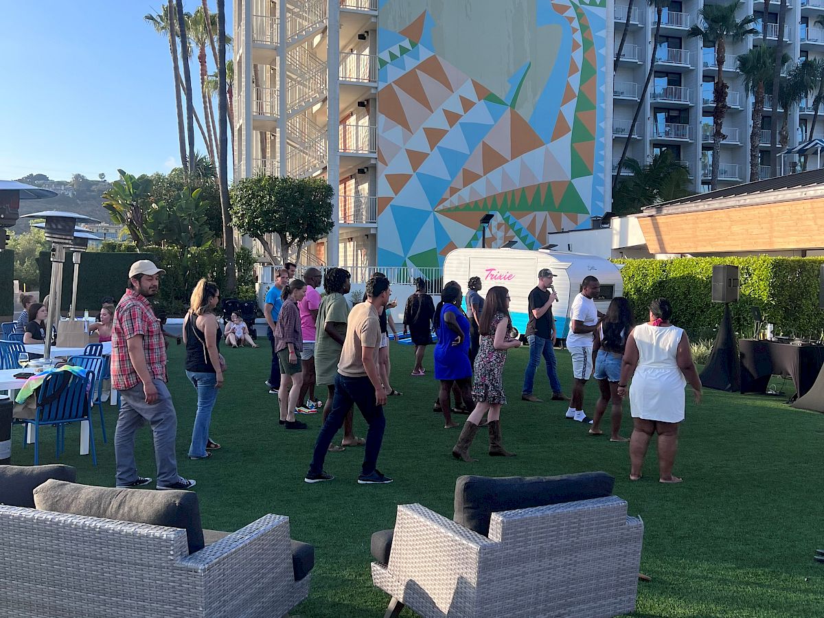 People are gathered on a grassy area with outdoor furniture, near a tall building with a colorful, geometric mural in the background.