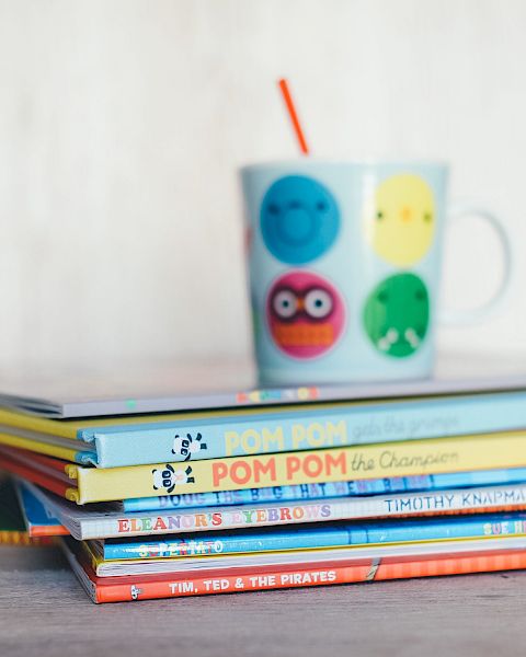 A stack of children's books with a colorful mug on top, featuring playful faces and a red straw, placed on a wooden surface.