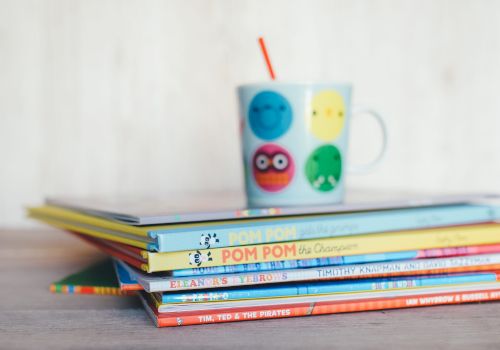 A stack of children's books with a colorful mug on top, featuring playful faces and a red straw, placed on a wooden surface.