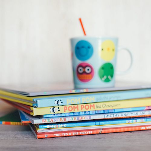 A stack of children's books with a colorful mug on top, featuring playful faces and a red straw, placed on a wooden surface.