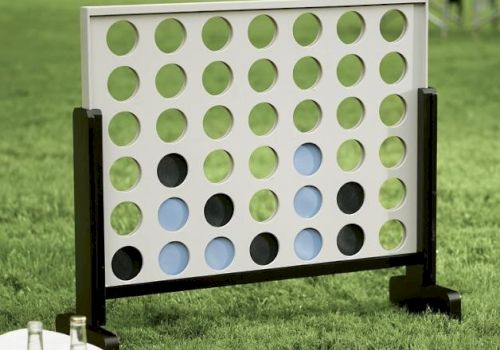 The image shows an outdoor Connect Four game set on grass, accompanied by drinks and a basket with plates.