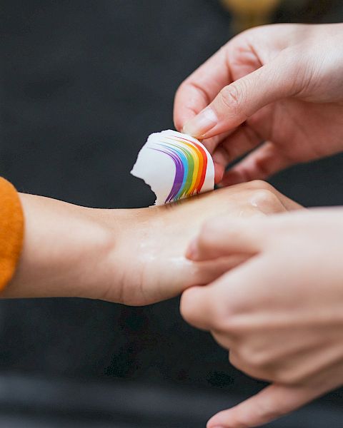 Two hands are seen, one person is applying a rainbow-colored bandage to the wrist of the other person’s hand, which is slightly outstretched.