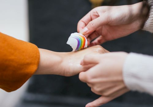 Two hands are seen, one person is applying a rainbow-colored bandage to the wrist of the other person’s hand, which is slightly outstretched.
