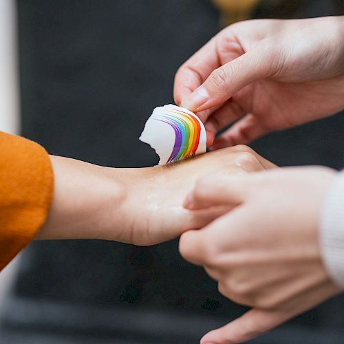 Two hands are seen, one person is applying a rainbow-colored bandage to the wrist of the other person’s hand, which is slightly outstretched.