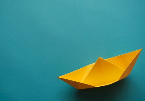 The image shows a yellow origami paper boat on a solid turquoise background.