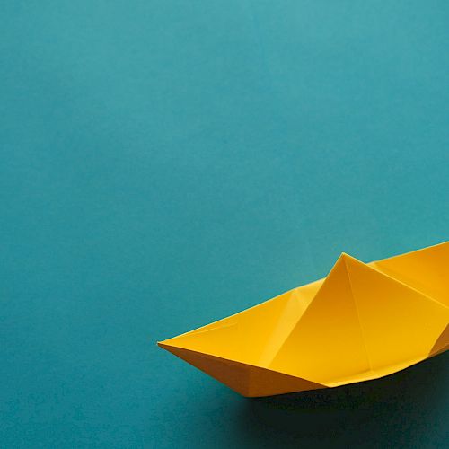 The image shows a yellow origami paper boat on a solid turquoise background.