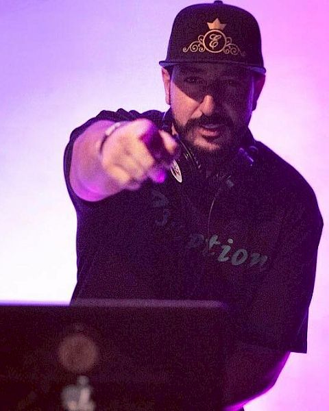 A DJ is performing on stage, wearing a hat and headset, pointing toward the camera, with a purple-lit background.