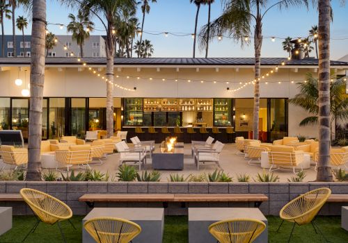 An outdoor lounge area with yellow chairs, string lights, and palm trees. There's a fire pit in the center and a bar in the background, ending the sentence.