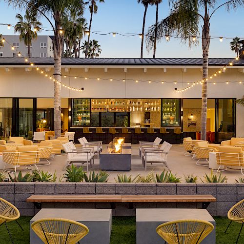 An outdoor lounge area with yellow chairs, string lights, and palm trees. There's a fire pit in the center and a bar in the background, ending the sentence.