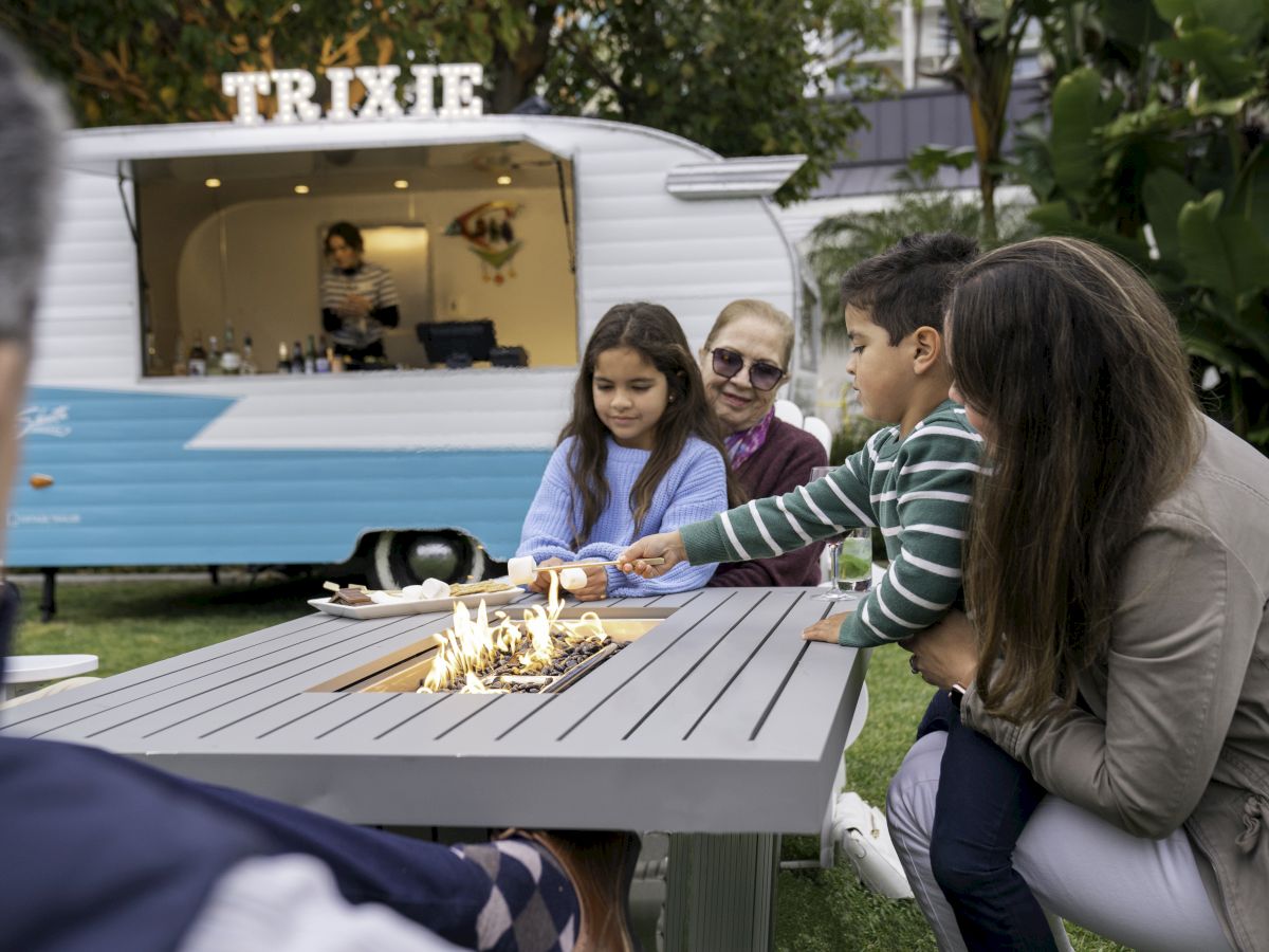 People are sitting around a table with a small fire on it, making s'mores. A food truck labeled 