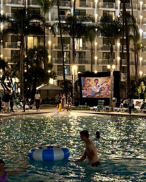 A nighttime pool scene with people swimming and relaxing, and a movie playing on a big screen in front of a lit-up building and palm trees.