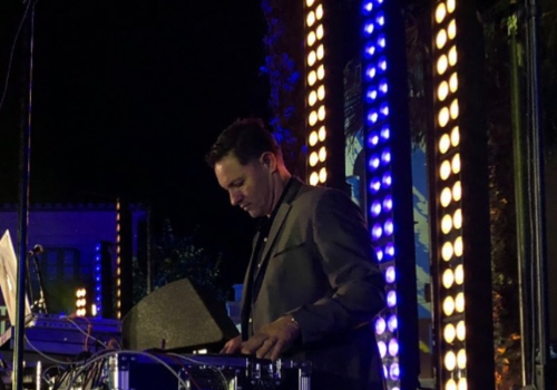 A man in a suit is DJing at an outdoor event with colorful, vertical lights and electronic equipment around him.