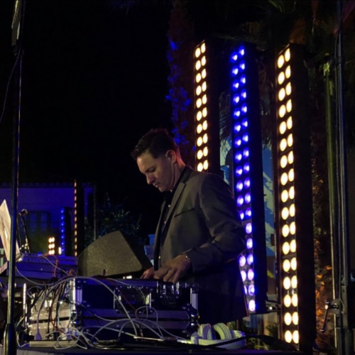 A man in a suit is DJing at an outdoor event with colorful, vertical lights and electronic equipment around him.