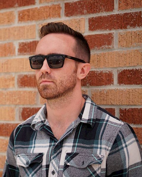 A man wearing sunglasses and a plaid shirt stands against a brick wall, looking off into the distance.