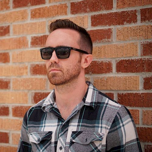 A man wearing sunglasses and a plaid shirt stands against a brick wall, looking off into the distance.