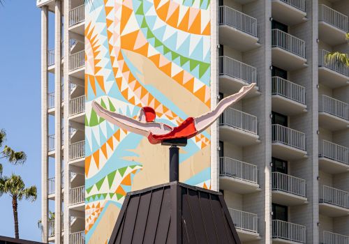 This image shows a tall building with a colorful geometric mural and a red sculpture on the roof of an adjacent structure, with some palm trees visible.