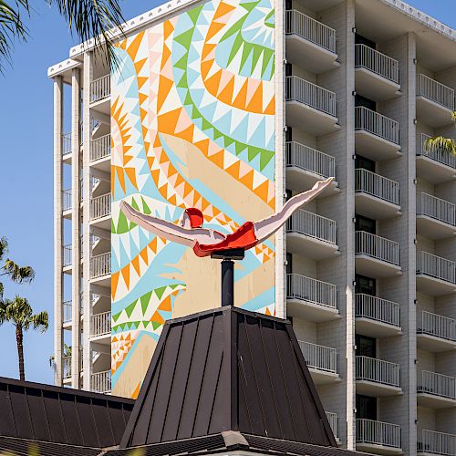 This image shows a tall building with a colorful geometric mural and a red sculpture on the roof of an adjacent structure, with some palm trees visible.