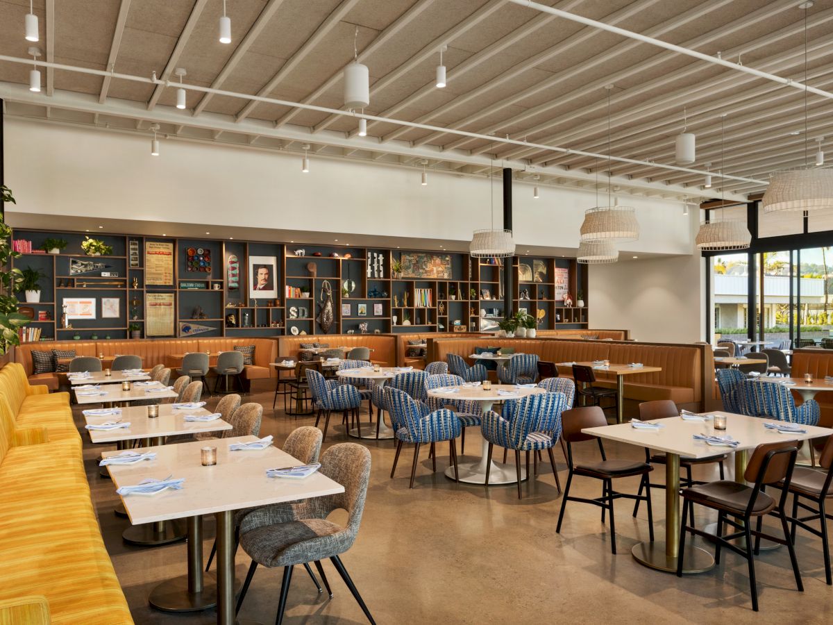 The image shows a modern, well-lit restaurant interior with yellow booth seating, various chairs, neatly set tables, and a large bookshelf against one wall.