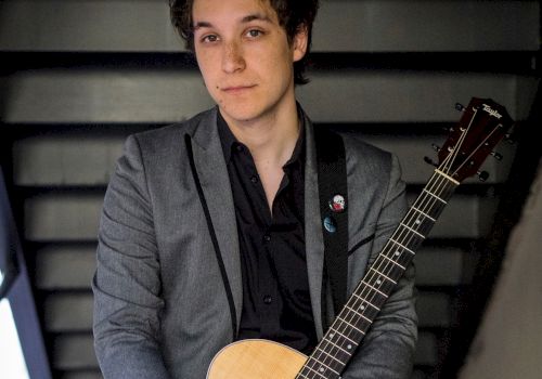 A person is standing on a staircase, holding an acoustic guitar, and is dressed in a gray blazer over a black shirt, looking at the camera.