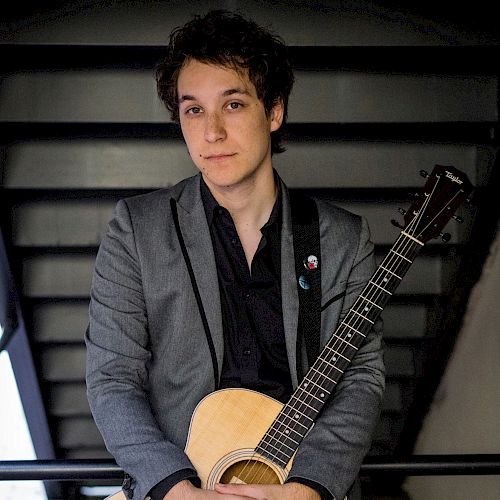 A person is standing on a staircase, holding an acoustic guitar, and is dressed in a gray blazer over a black shirt, looking at the camera.