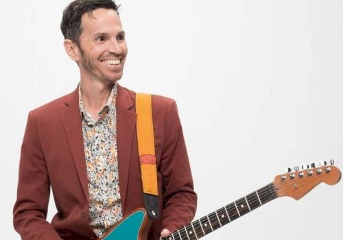 A smiling man in a red blazer is holding a turquoise electric guitar, standing against a plain white background.