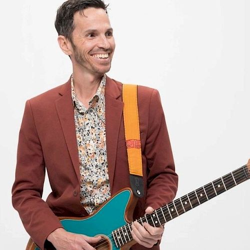 A smiling man in a red blazer is holding a turquoise electric guitar, standing against a plain white background.