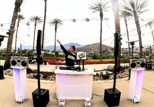 A DJ is performing outdoors in a scenic location with palm trees and mountains in the background, surrounded by sound equipment and colorful decorations.