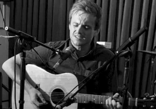 A person is playing an acoustic guitar while singing into a microphone, with a drum set visible in the background.