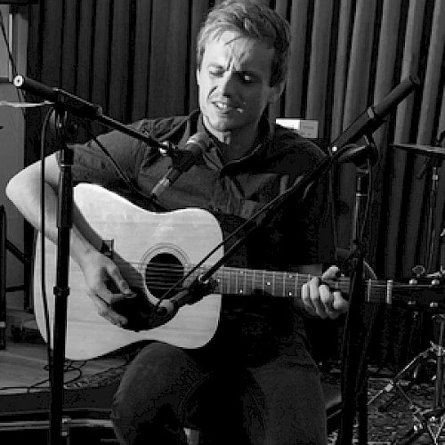 A person is playing an acoustic guitar while singing into a microphone, with a drum set visible in the background.