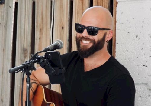 A person with a beard and sunglasses is playing an acoustic guitar and smiling, likely performing music outdoors.